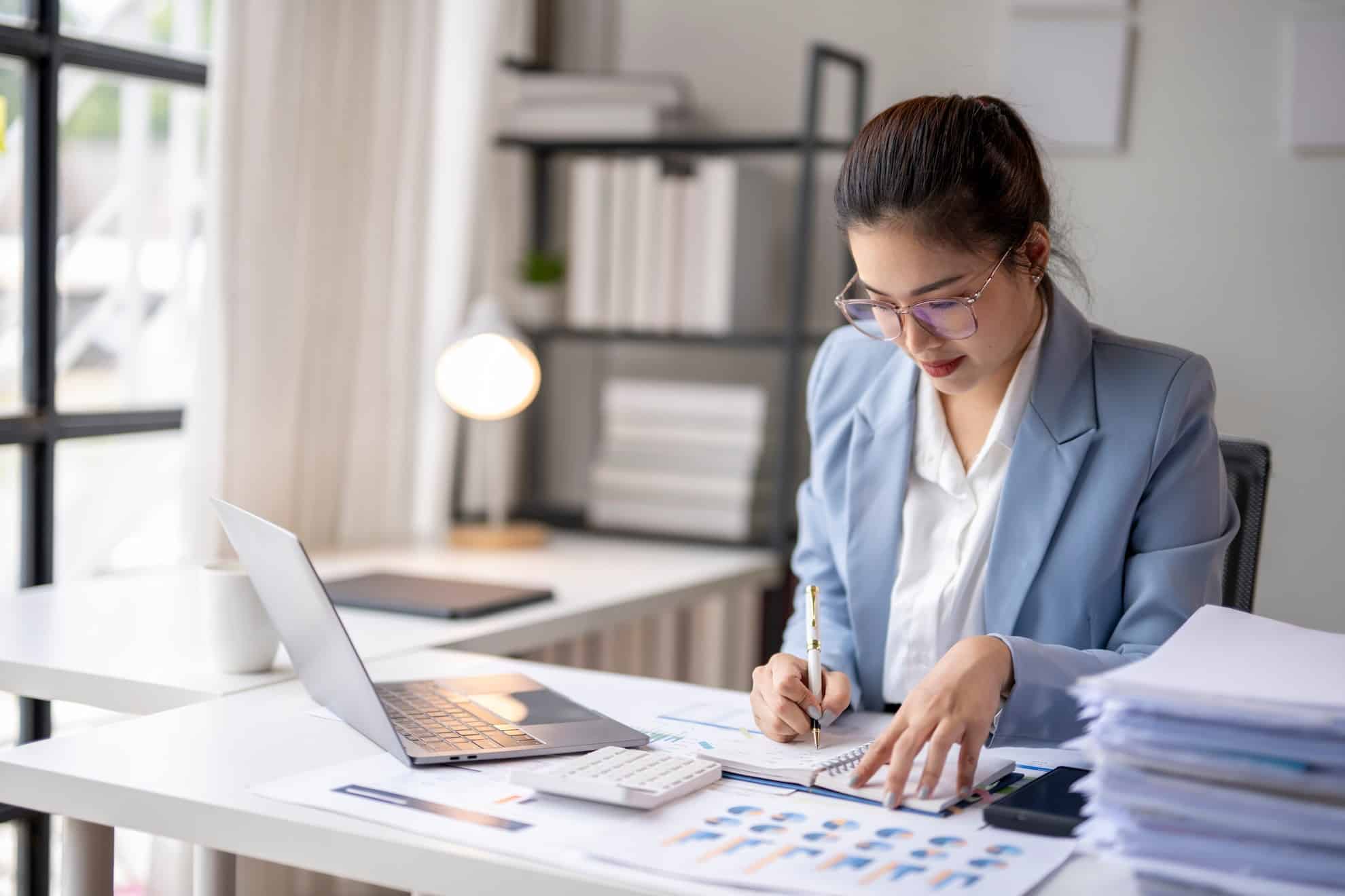 A female business professional reviewing financial reports and accounting data at her desk with a laptop.