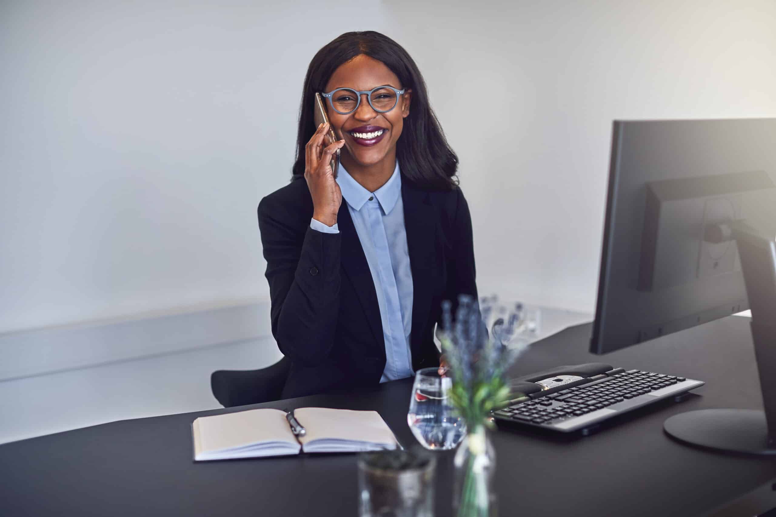 A female tax accountant holding her phone to her ear in an office with a notepad and a computer at the desk.