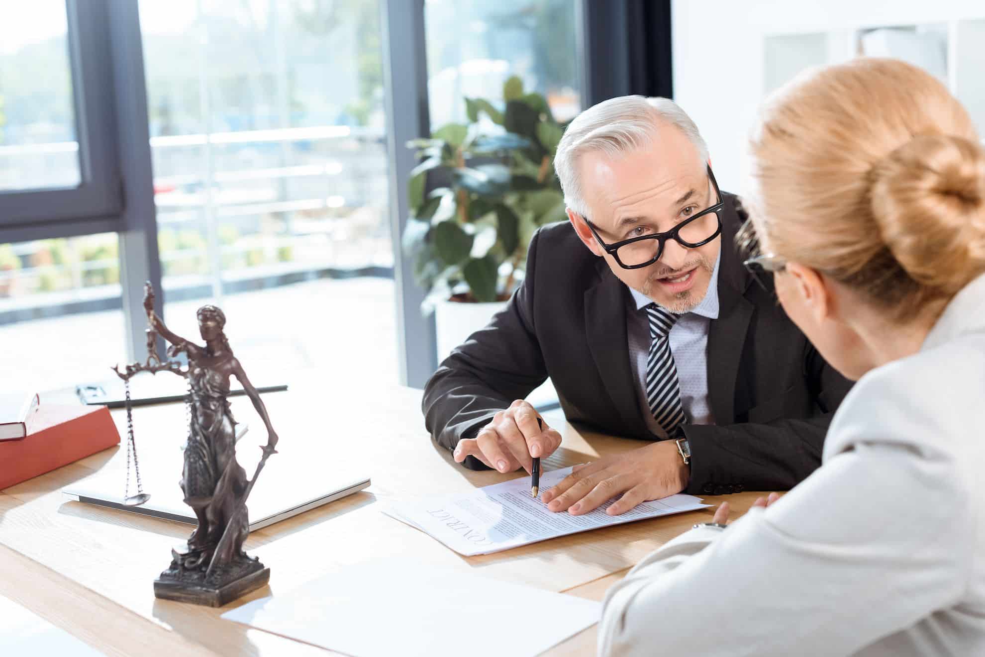 A lawyer explains legal terms of a power of attorney document to a female client at a modern office desk, with a statue of Lady Justice in the foreground.